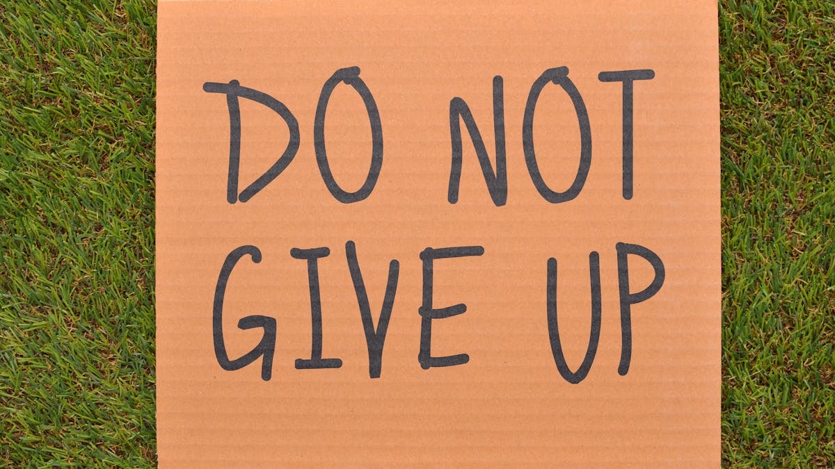 Cardboard sign reading 'Do Not Give Up' placed on green turf, symbolizing perseverance.